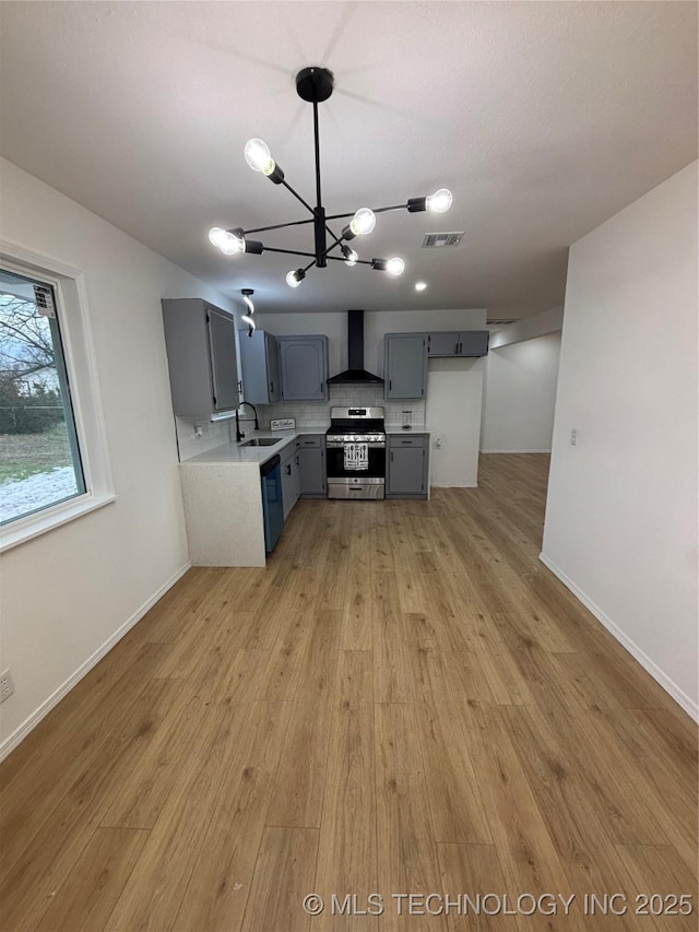 kitchen with a notable chandelier, wall chimney range hood, gray cabinets, light wood-type flooring, and appliances with stainless steel finishes