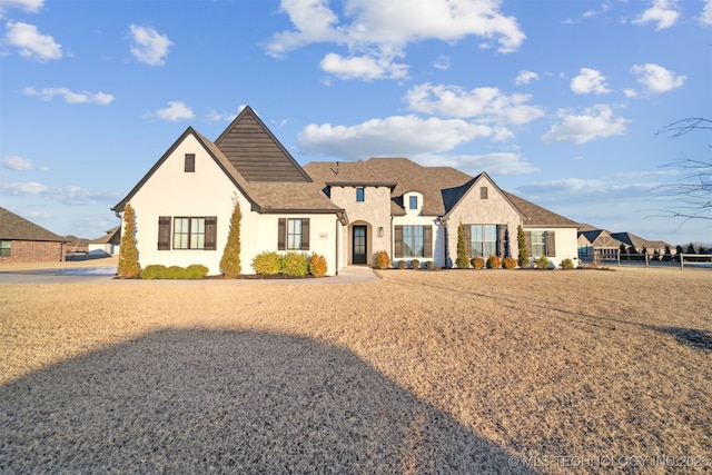 french provincial home featuring stucco siding, roof with shingles, and fence