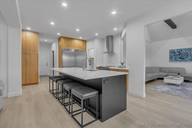 kitchen featuring wall chimney exhaust hood, stainless steel built in fridge, an island with sink, a breakfast bar area, and sink