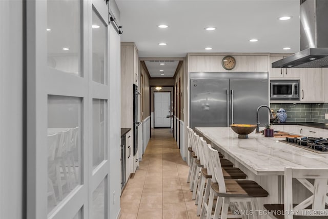 kitchen featuring light tile patterned floors, a kitchen breakfast bar, built in appliances, light stone counters, and exhaust hood