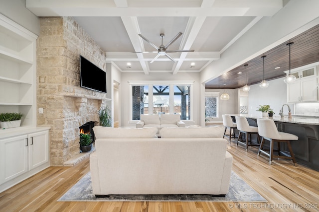 living room featuring ceiling fan, coffered ceiling, a stone fireplace, built in features, and light wood-type flooring