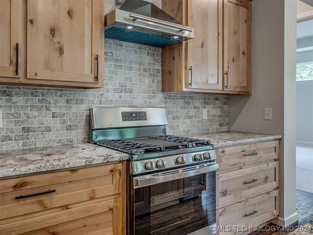 kitchen with light stone countertops, light brown cabinets, wall chimney range hood, stainless steel gas range, and tasteful backsplash