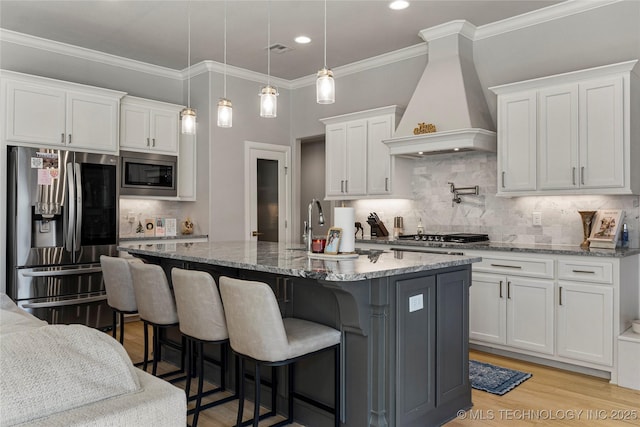 kitchen with appliances with stainless steel finishes, light stone counters, custom range hood, a kitchen island with sink, and white cabinets