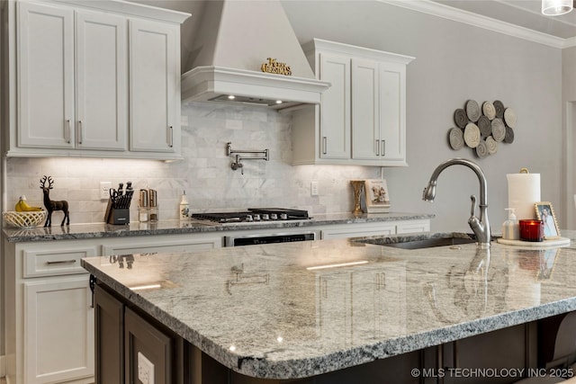 kitchen with premium range hood, stainless steel gas cooktop, a kitchen island with sink, sink, and white cabinetry