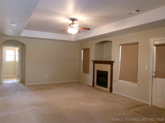 unfurnished living room featuring a tiled fireplace, ceiling fan, a tray ceiling, and light colored carpet