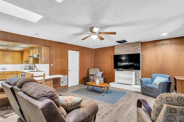 living room featuring wooden walls, a textured ceiling, ceiling fan, light hardwood / wood-style floors, and a skylight