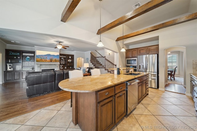 kitchen with a kitchen island with sink, stainless steel appliances, beamed ceiling, light tile patterned floors, and ceiling fan