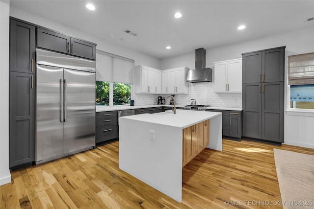 kitchen with stainless steel appliances, white cabinets, light hardwood / wood-style floors, wall chimney range hood, and a kitchen island with sink