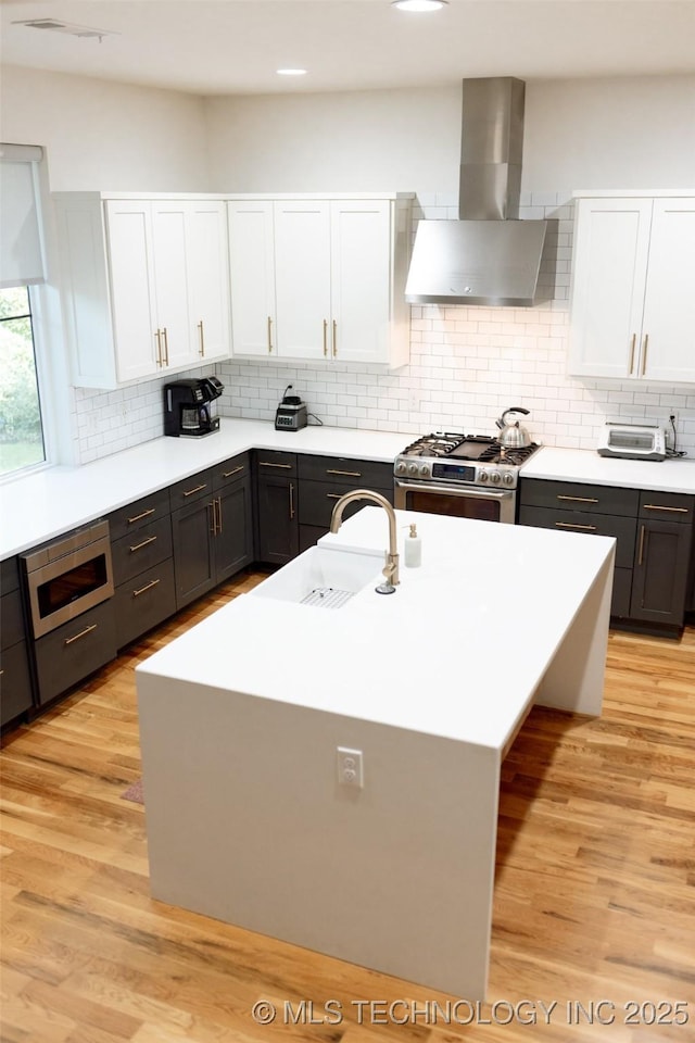 kitchen featuring an island with sink, wall chimney range hood, backsplash, white cabinets, and appliances with stainless steel finishes