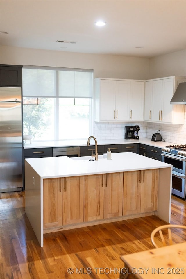 kitchen featuring white cabinetry, hardwood / wood-style flooring, a center island with sink, wall chimney range hood, and appliances with stainless steel finishes