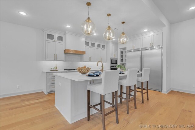 kitchen with a kitchen island with sink, built in appliances, premium range hood, pendant lighting, and white cabinets