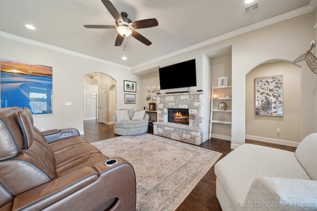 living room with ceiling fan, dark hardwood / wood-style flooring, built in features, ornamental molding, and a stone fireplace