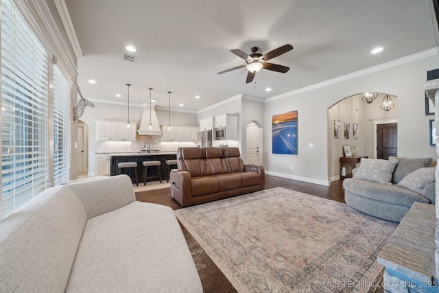 living room with ceiling fan with notable chandelier, sink, ornamental molding, and dark wood-type flooring