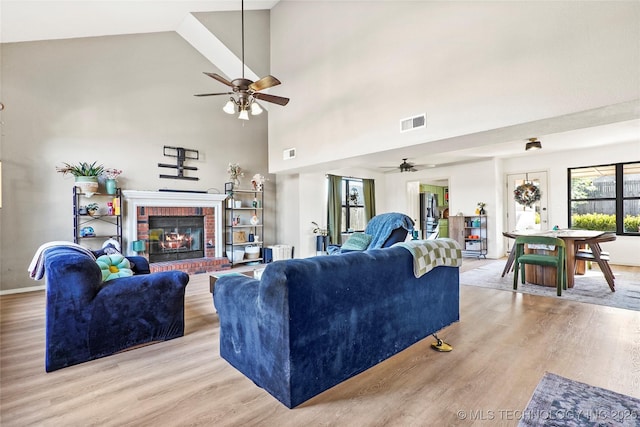 living room featuring high vaulted ceiling, a brick fireplace, ceiling fan, and light wood-type flooring