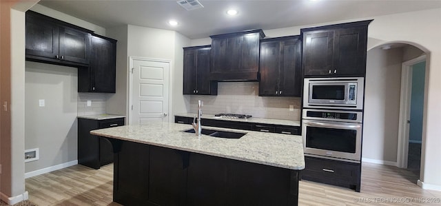 kitchen with appliances with stainless steel finishes, decorative backsplash, a kitchen island with sink, light wood-type flooring, and light stone counters