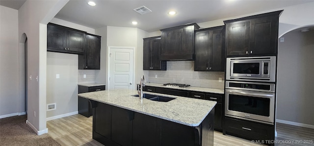 kitchen featuring an island with sink, stainless steel appliances, backsplash, light stone counters, and sink