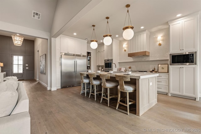 kitchen featuring a kitchen bar, built in appliances, white cabinetry, hanging light fixtures, and an island with sink