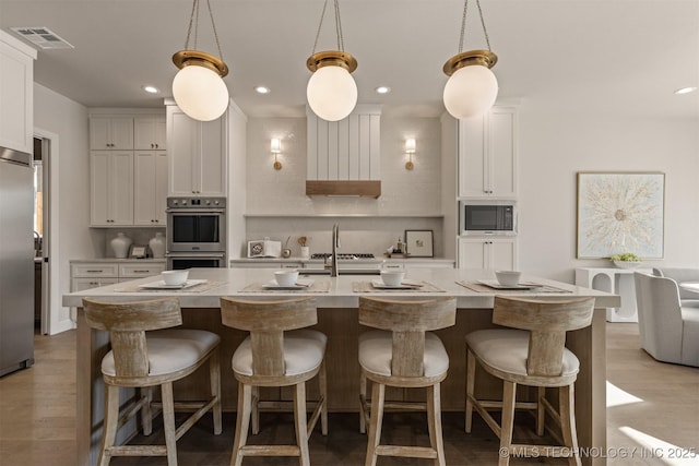 kitchen with decorative light fixtures, white cabinetry, a kitchen island with sink, appliances with stainless steel finishes, and light stone counters