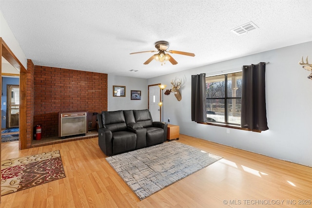 living room with ceiling fan, a brick fireplace, light wood-type flooring, and a textured ceiling