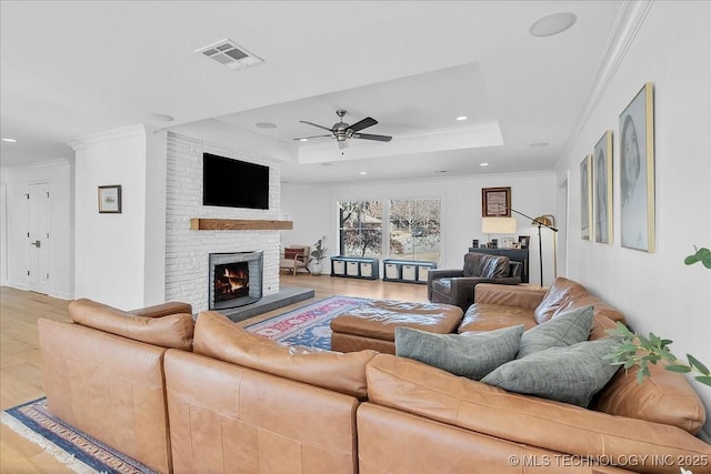 living room with ceiling fan, a raised ceiling, a fireplace, crown molding, and light hardwood / wood-style flooring