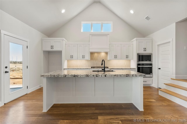 kitchen with sink, light stone counters, an island with sink, white cabinets, and stainless steel oven