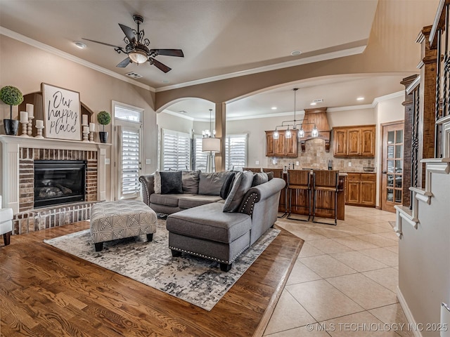 living room featuring a fireplace, light tile patterned flooring, ceiling fan with notable chandelier, and ornamental molding