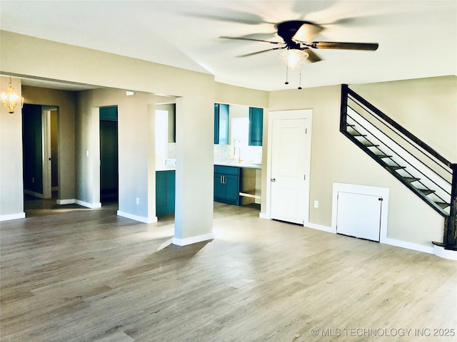 unfurnished living room featuring baseboards, stairway, wood finished floors, a sink, and ceiling fan with notable chandelier