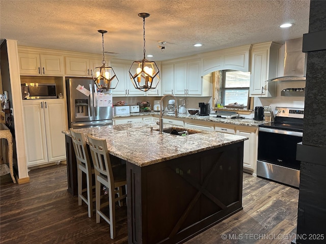 kitchen with wall chimney exhaust hood, stainless steel appliances, sink, hanging light fixtures, and a center island with sink