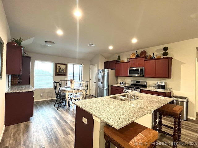 kitchen with an island with sink, a kitchen breakfast bar, stainless steel appliances, light stone countertops, and dark wood-type flooring