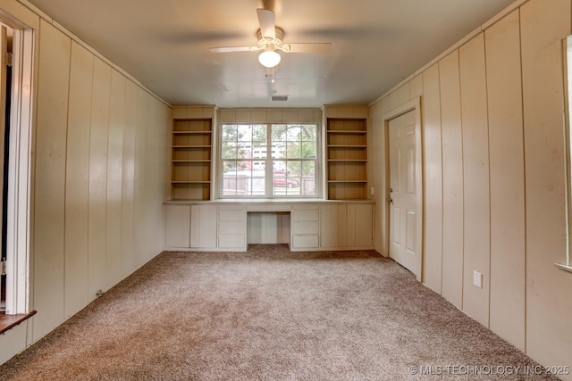 unfurnished office featuring ceiling fan, built in desk, light colored carpet, and wood walls