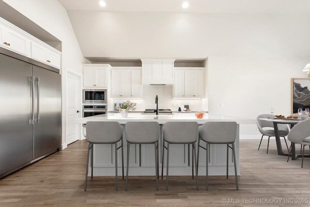 kitchen with wood finished floors, built in appliances, a kitchen island with sink, white cabinetry, and backsplash