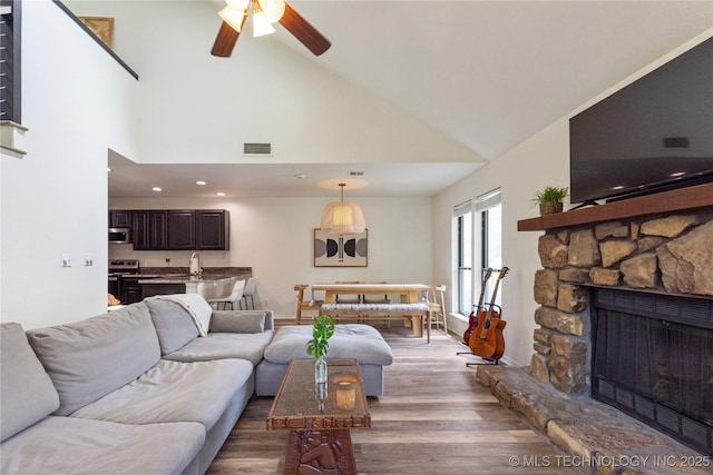 living room with ceiling fan, sink, dark wood-type flooring, a stone fireplace, and high vaulted ceiling