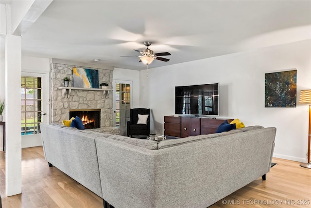 living room with a ceiling fan, a stone fireplace, baseboards, and wood finished floors