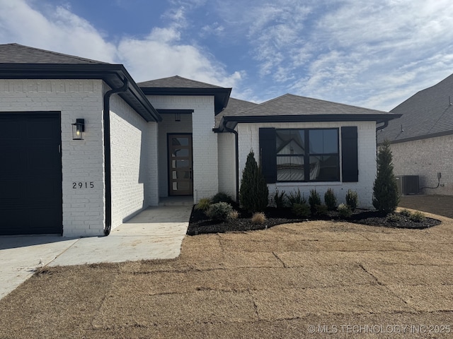 view of front of house featuring central air condition unit, an attached garage, a shingled roof, and brick siding