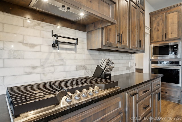 kitchen with dark wood-type flooring, premium range hood, backsplash, and stainless steel appliances