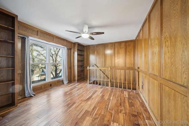 empty room with ceiling fan, built in shelves, wood-type flooring, and wood walls