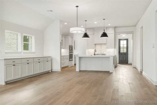 kitchen featuring white cabinetry, backsplash, hanging light fixtures, a center island with sink, and light hardwood / wood-style flooring