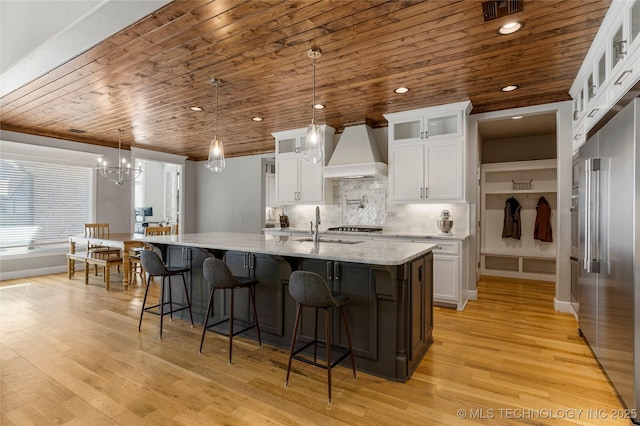 kitchen featuring high quality fridge, decorative light fixtures, white cabinetry, a kitchen island with sink, and custom range hood