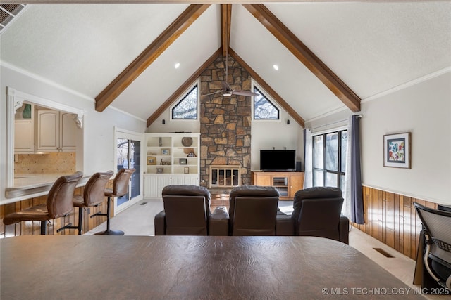 living room with ceiling fan, wooden walls, a stone fireplace, and vaulted ceiling with beams