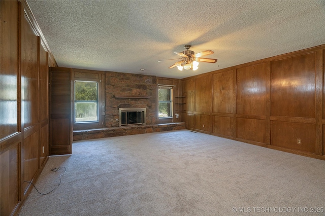 unfurnished living room with ceiling fan, light colored carpet, wood walls, and a stone fireplace