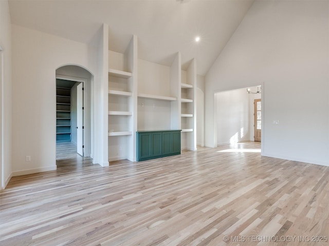 unfurnished living room with light wood-type flooring, high vaulted ceiling, built in features, arched walkways, and baseboards