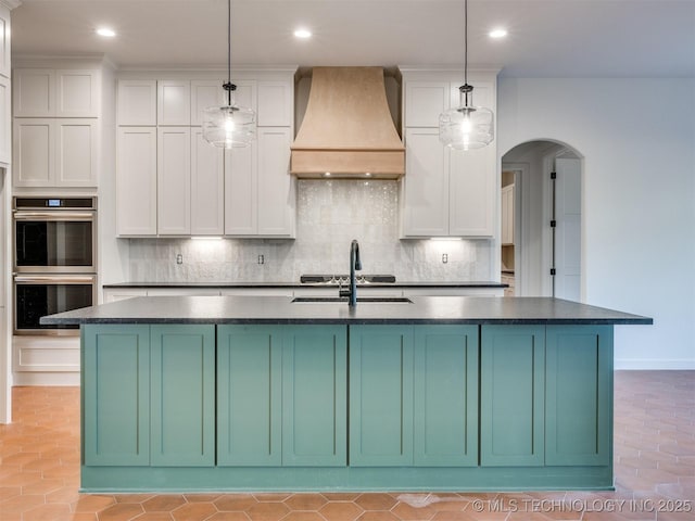 kitchen with tasteful backsplash, premium range hood, arched walkways, stainless steel double oven, and a kitchen island with sink