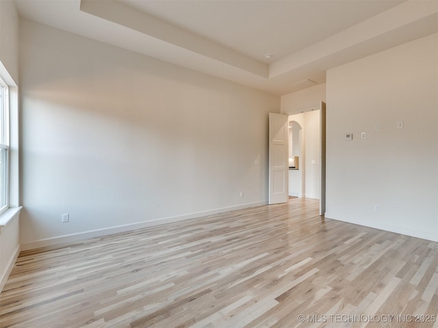 empty room with a raised ceiling, baseboards, arched walkways, and light wood finished floors