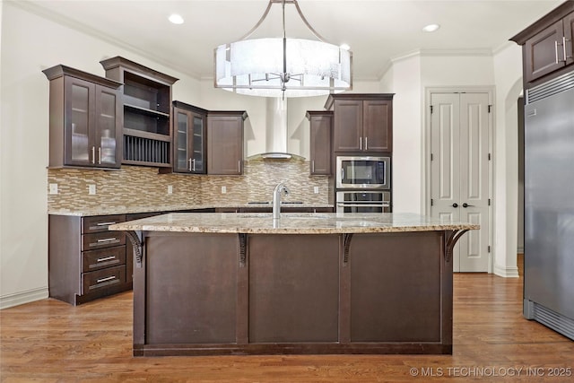 kitchen featuring light stone countertops, wall chimney exhaust hood, a kitchen bar, built in appliances, and dark brown cabinets