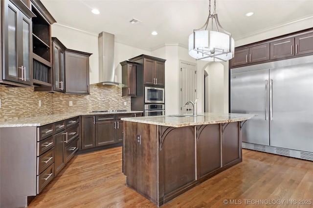 kitchen with sink, tasteful backsplash, a kitchen island with sink, wall chimney range hood, and built in appliances