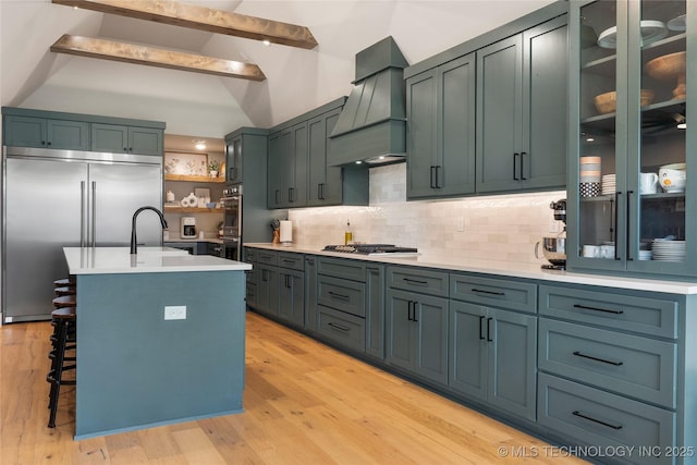 kitchen featuring tasteful backsplash, sink, a kitchen island with sink, stainless steel appliances, and custom range hood