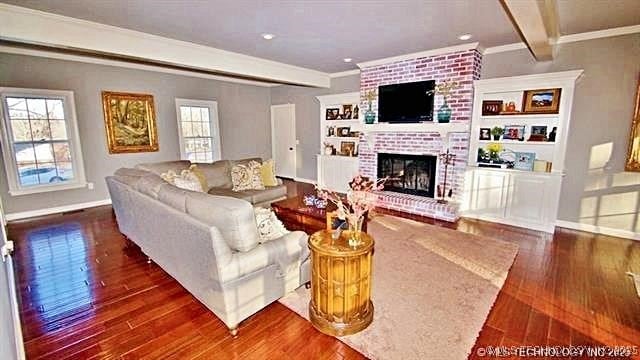 living room featuring a brick fireplace, dark wood-type flooring, crown molding, and beamed ceiling