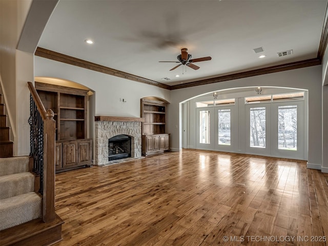 unfurnished living room with crown molding, ceiling fan, wood-type flooring, a fireplace, and built in shelves