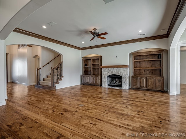 unfurnished living room with built in shelves, a stone fireplace, wood-type flooring, ornamental molding, and ceiling fan