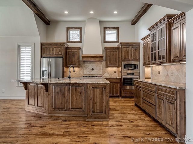 kitchen with stainless steel appliances, premium range hood, a center island with sink, and beam ceiling